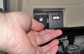 A hand opening a black hood release switch, which is right next to the fuel door switch