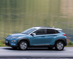 A blue Kona Electric driving in front of a body of water